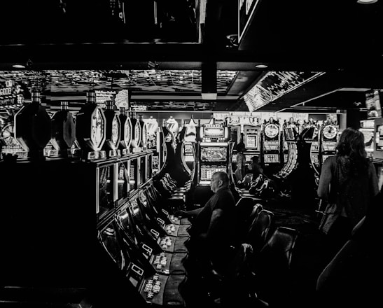 A dimly lit casino filled with rows of slot machines, some occupied by people. The machines' lights contrast with the dark surroundings, creating a vibrant yet subdued atmosphere.