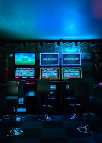 Dimly lit gaming arcade featuring a variety of brightly illuminated slot machines and video gaming screens. Neon signs and colorful displays create a vibrant atmosphere. The arrangement of gaming machines suggests an inviting space for entertainment, with chairs positioned for players.