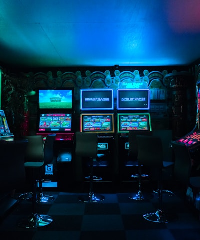 Dimly lit gaming arcade featuring a variety of brightly illuminated slot machines and video gaming screens. Neon signs and colorful displays create a vibrant atmosphere. The arrangement of gaming machines suggests an inviting space for entertainment, with chairs positioned for players.