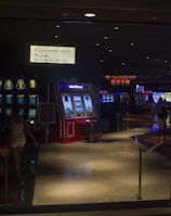 A dimly lit casino with several rows of slot machines, some displaying bright graphics and lights. There are a few people scattered throughout the area, engaging with the machines. The floor is reflective and polished, adding to the ambiance, while the ceiling is dotted with numerous small lights.
