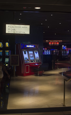 A dimly lit casino with several rows of slot machines, some displaying bright graphics and lights. There are a few people scattered throughout the area, engaging with the machines. The floor is reflective and polished, adding to the ambiance, while the ceiling is dotted with numerous small lights.