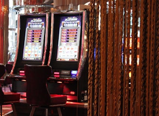 A pair of casino slot machines, each with detailed screens displaying various game outcomes and jackpot amounts. The chairs in front of the slot machines are dark and cushioned for comfort. The background features a hanging curtain made of golden ropes and other casino decorations are visible.