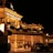 A luxurious casino at night, illuminated with warm, golden lights. The building features elegant architecture with columns and decorative elements. A few people are walking towards the entrance, and there are a couple of parked cars nearby.