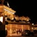 A luxurious casino at night, illuminated with warm, golden lights. The building features elegant architecture with columns and decorative elements. A few people are walking towards the entrance, and there are a couple of parked cars nearby.