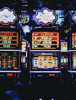 A row of colorful slot machines lit with bright neon lights, showcasing various graphical elements on the screens, indicating different games and jackpots. The atmosphere suggests a lively casino setting, bustling with excitement.