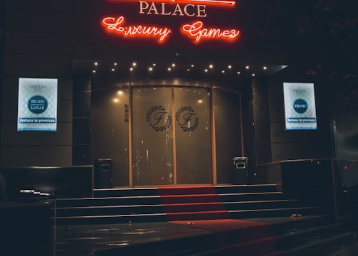 A neon-lit casino entrance with a red carpet leading up to large double doors. The sign reads 'Fortuna Palace Luxury Games' in bright red and white neon lights. The dimly lit atmosphere suggests a night-time setting. There are two digital displays on either side of the entrance showing promotional content.