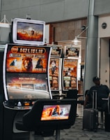 A collection of slot machines is situated inside a building with bright, colorful screens displaying various games and gaming themes. A person is seated nearby, seemingly relaxed with luggage beside them, and the atmosphere is lively with electronic displays.