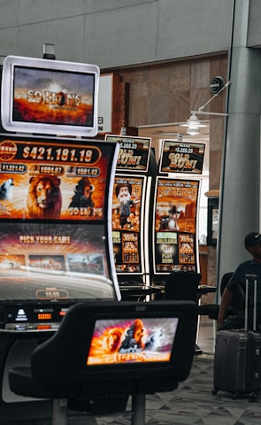 A collection of slot machines is situated inside a building with bright, colorful screens displaying various games and gaming themes. A person is seated nearby, seemingly relaxed with luggage beside them, and the atmosphere is lively with electronic displays.